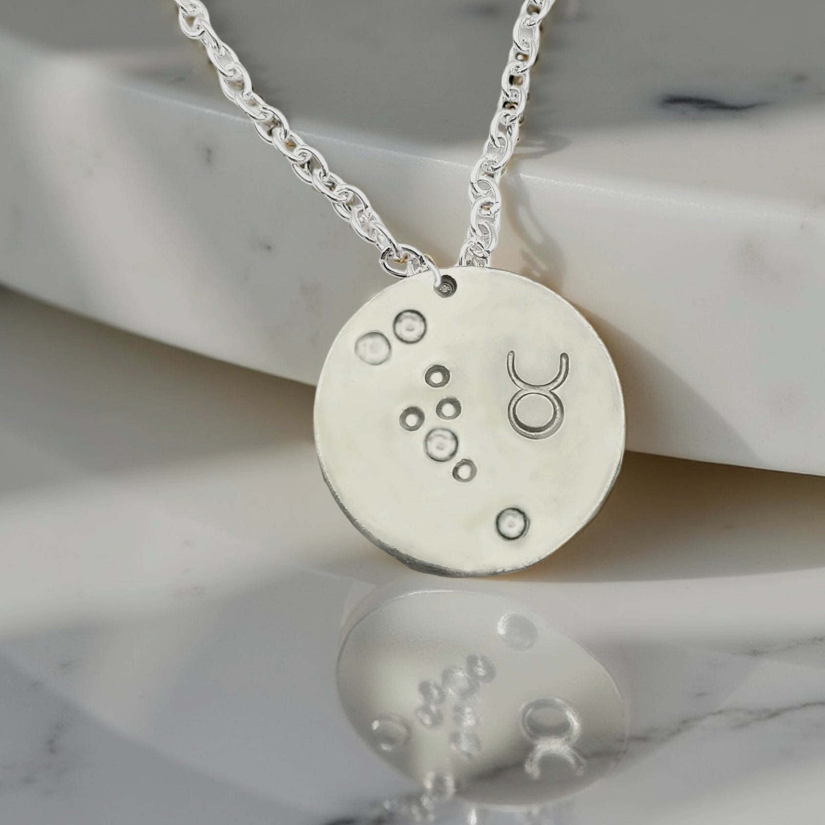 April Taurus  necklace pendant with hand set diamond colored glass rhinestones forming the constellation of Taurus. Hand stamped with the zodiac symbol for Taurus, on a 3/4th of an inch silver toned circle hanging from a silver toned chain.