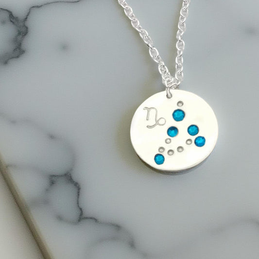 December Capricorn necklace pendant with hand set tanzanite colored glass rhinestones forming the constellation of Capricorn. Hand stamped with the zodiac symbol for Capricorn, on a 3/4th of an inch silver toned circle hanging from a silver toned chain.