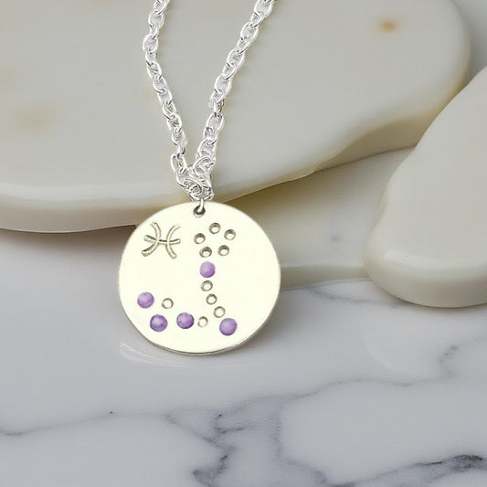 February Pisces necklace pendant with hand set amethyst colored glass rhinestones forming the constellation of Pisces. Hand stamped with the zodiac symbol for Pisces, on a 3/4th of an inch silver toned circle hanging from a silver toned chain.