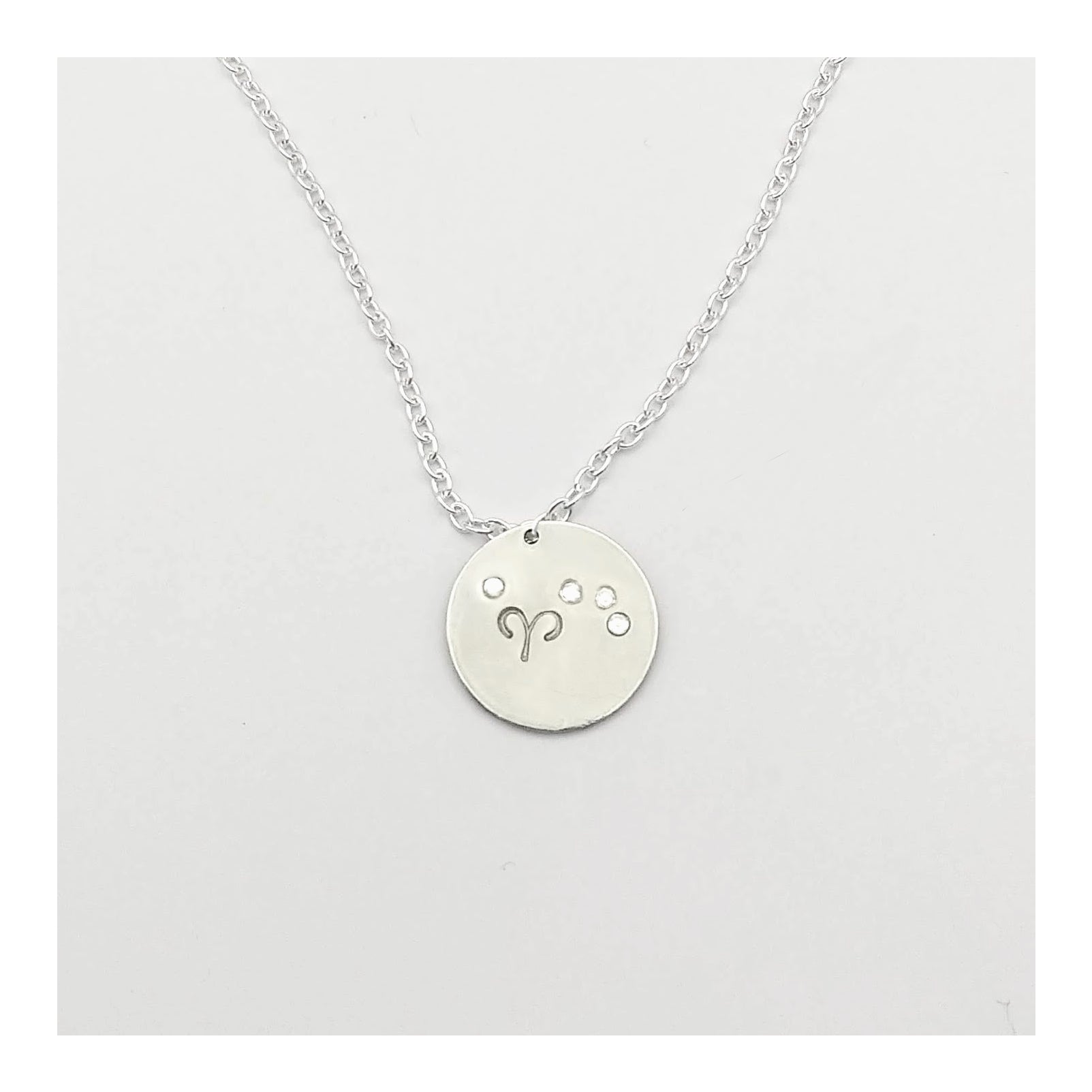 April Aries necklace pendant with hand set diamond colored glass rhinestones forming the constellation of Aries. Hand stamped with the zodiac symbol for aries, on a 3/4th of an inch silver toned circle hanging from a silver toned chain.