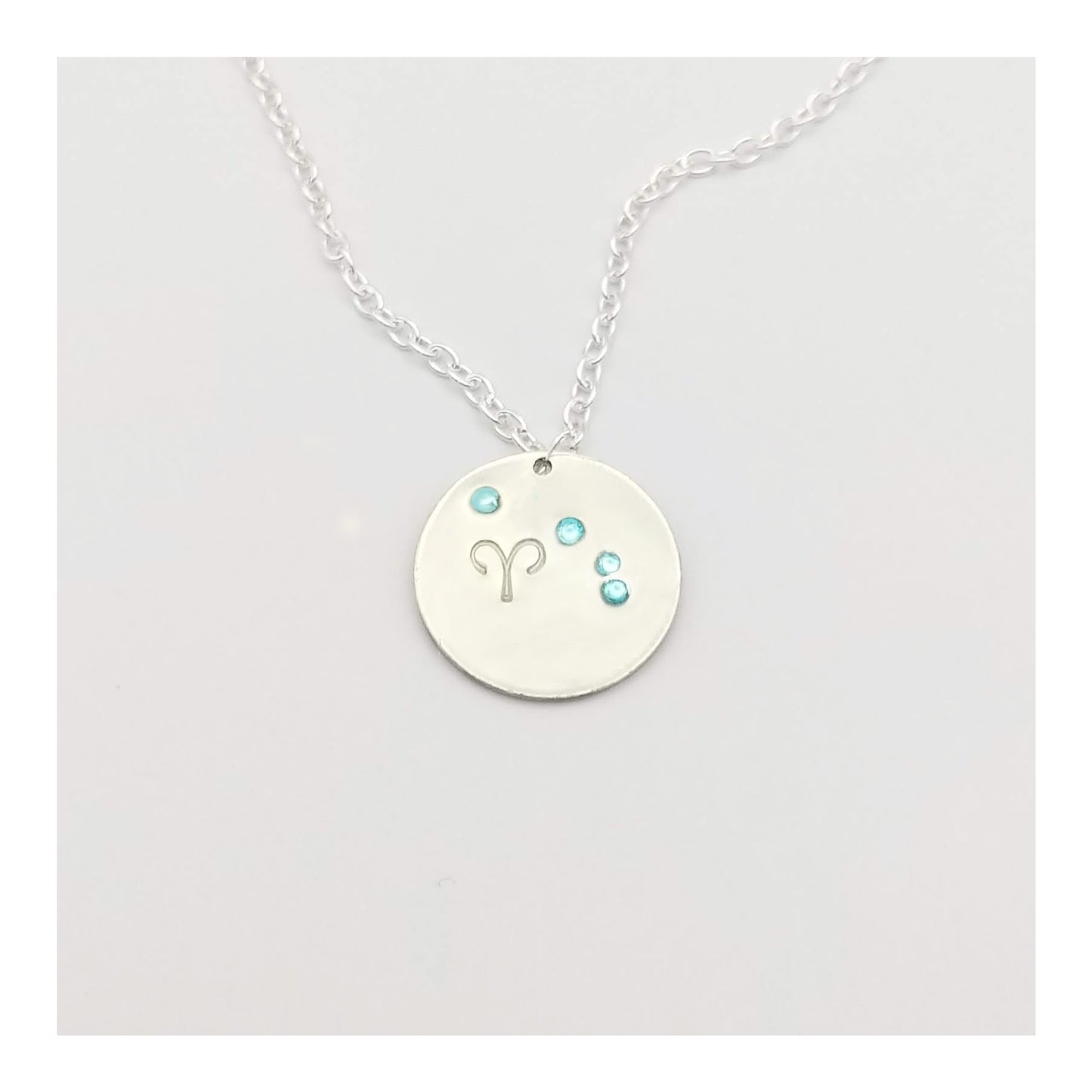 March Aries necklace pendant with hand set aquamarine colored glass rhinestones forming the constellation of Aries. Hand stamped with the zodiac symbol for aries, on a 3/4th of an inch silver toned circle hanging from a silver toned chain.