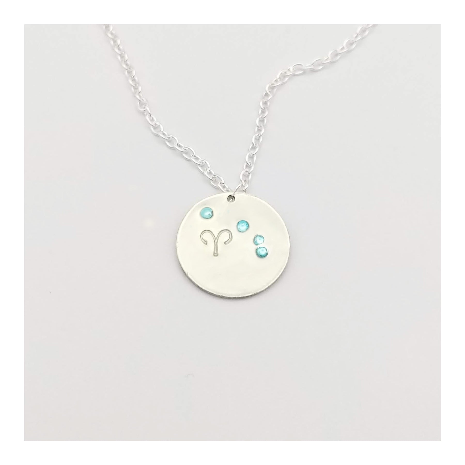 March Aries necklace pendant with hand set aquamarine colored glass rhinestones forming the constellation of Aries. Hand stamped with the zodiac symbol for aries, on a 3/4th of an inch silver toned circle hanging from a silver toned chain.