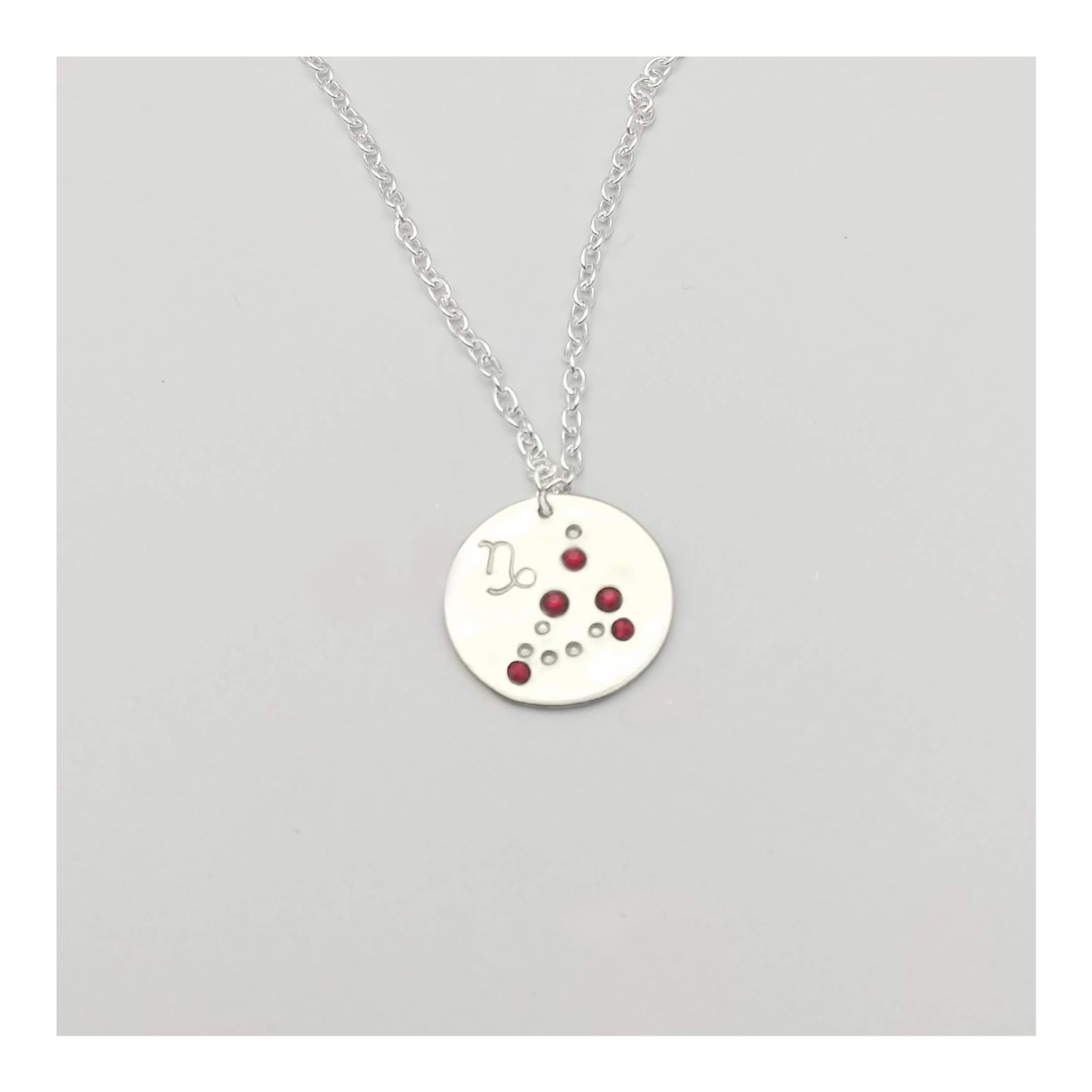 January Capricorn necklace pendant with hand set garnet colored glass rhinestones forming the constellation of Capricorn. Hand stamped with the zodiac symbol for Capricorn, on a 3/4th of an inch silver toned circle hanging from a silver toned chain.