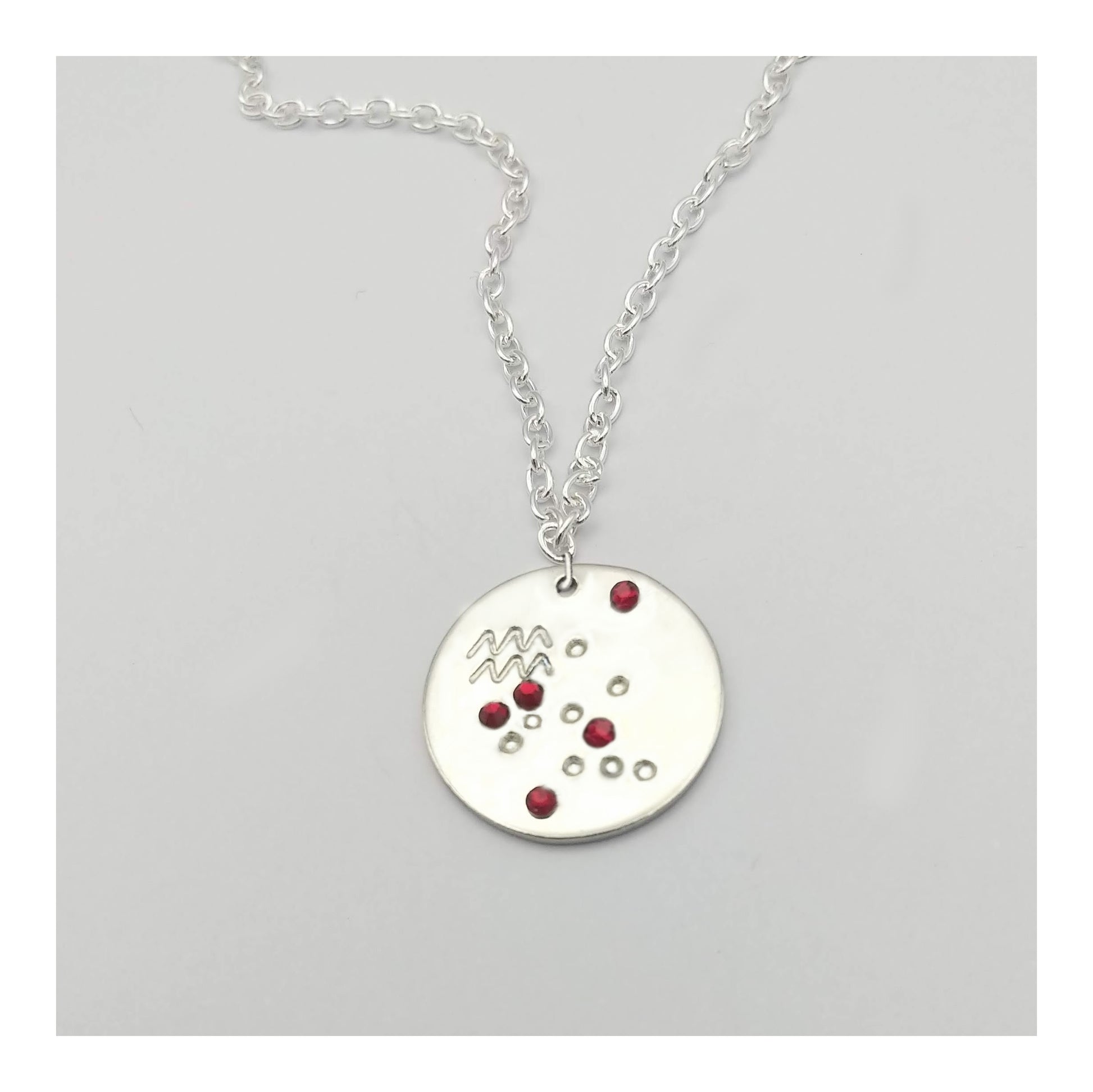 January Aquarius necklace pendant with hand set garnet colored glass rhinestones forming the constellation of Aquarius. Hand stamped with the zodiac symbol for Aquarius, on a 3/4th of an inch silver toned circle hanging from a silver toned chain.