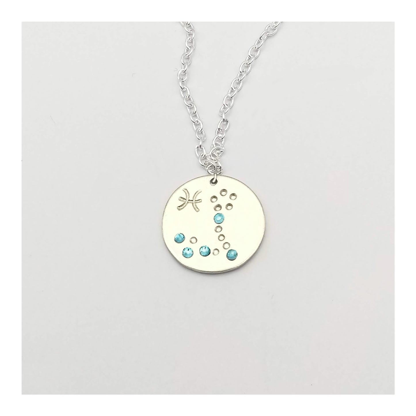 March Pisces necklace pendant with hand set aquamarine colored glass rhinestones forming the constellation of Pisces. Hand stamped with the zodiac symbol for Pisces, on a 3/4th of an inch silver toned circle hanging from a silver toned chain.