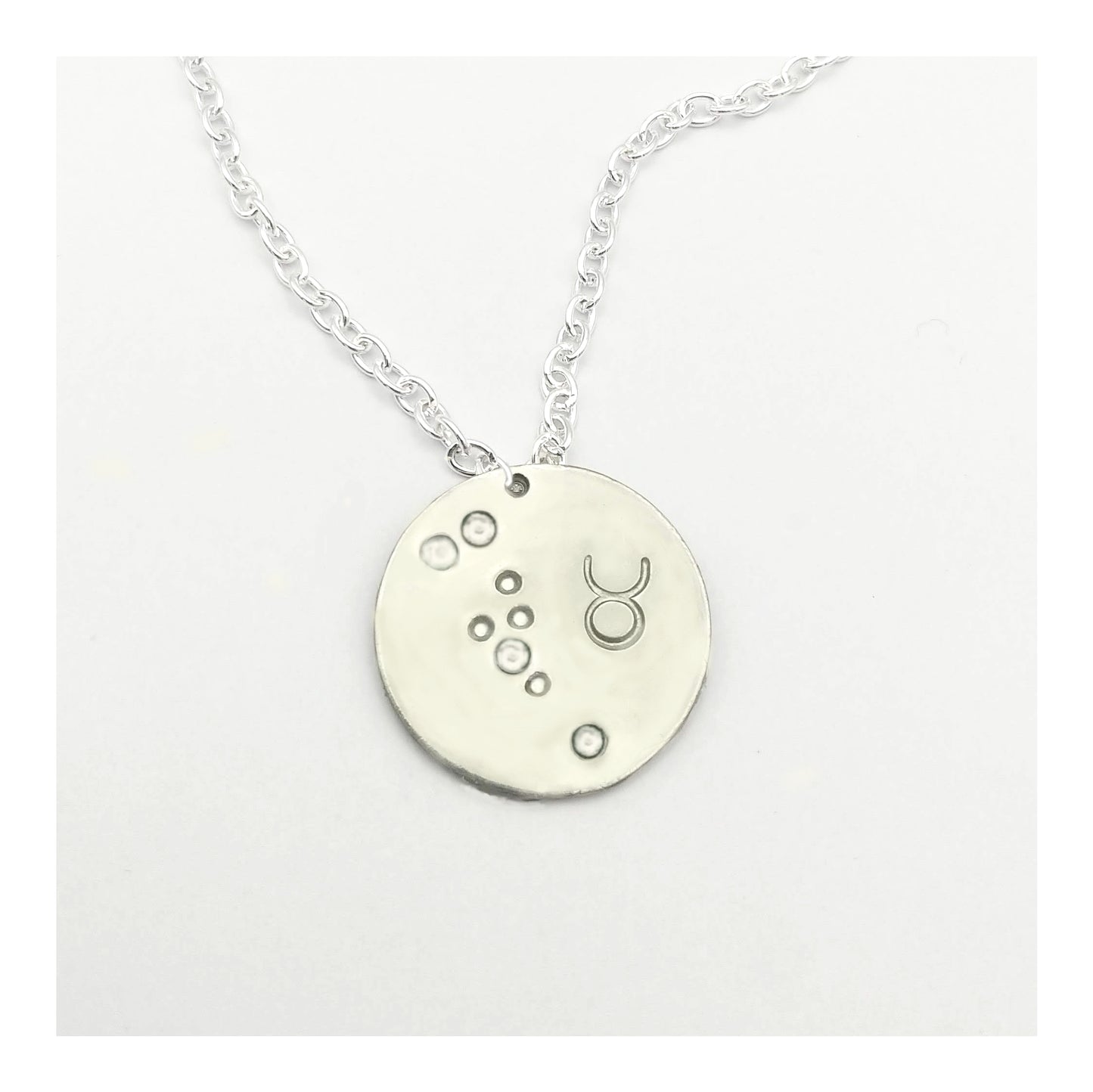 April Taurus  necklace pendant with hand set diamond colored glass rhinestones forming the constellation of Taurus. Hand stamped with the zodiac symbol for Taurus, on a 3/4th of an inch silver toned circle hanging from a silver toned chain.