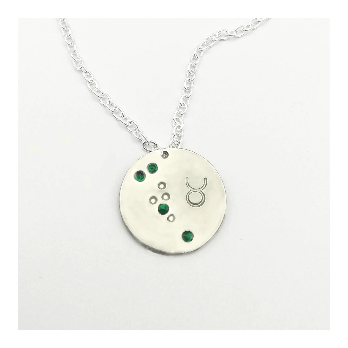 May Taurus necklace pendant with hand set emerald colored glass rhinestones forming the constellation of Taurus. Hand stamped with the zodiac symbol for Taurus, on a 3/4th of an inch silver toned circle hanging from a silver toned chain.