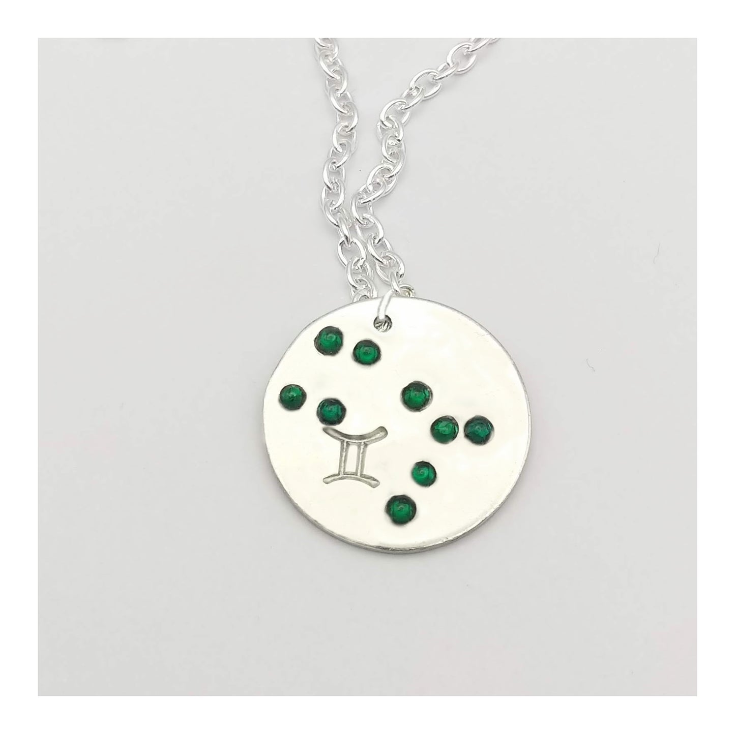 May Gemini necklace pendant with hand set emerald colored glass rhinestones forming the constellation of Gemini. Hand stamped with the zodiac symbol for Gemini, on a 3/4th of an inch silver toned circle hanging from a silver toned chain.