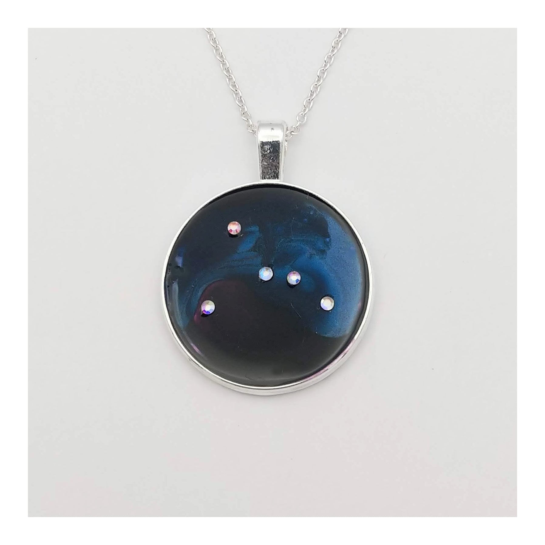 Dark blue with iridescent sparkling swirls of pinks, purples, silver, and monochromatic shades of blue enamel. With handset Aurora Borealis rhinestones to represent the star of the constellation of Cancer. The pendant is 1 inch silver toned with a 1/4th inch bail on a silver toned chain.