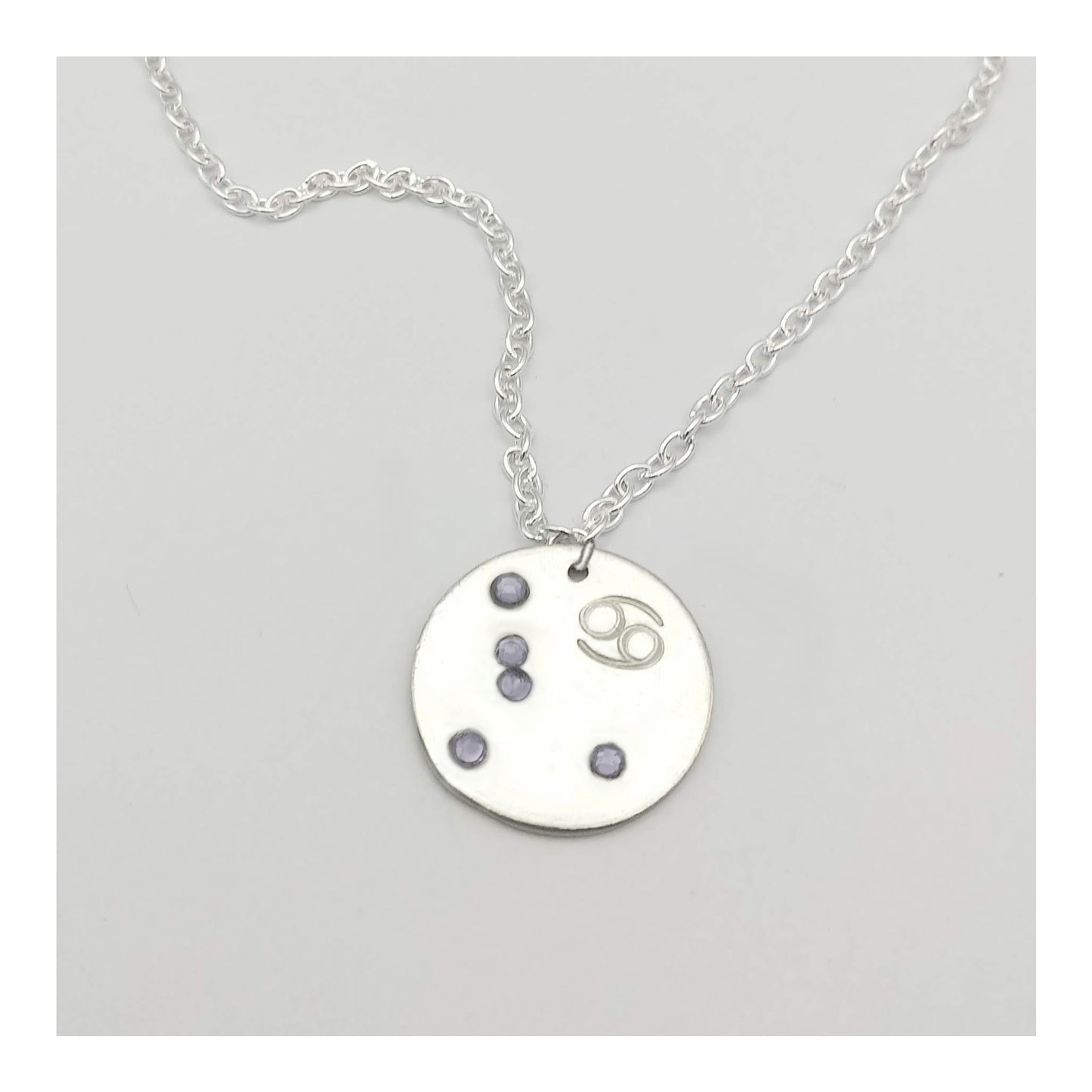 June Cancer necklace pendant with hand set alexandrite colored glass rhinestones forming the constellation of Cancer. Hand stamped with the zodiac symbol for Cancer, on a 3/4th of an inch silver toned circle hanging from a silver toned chain.