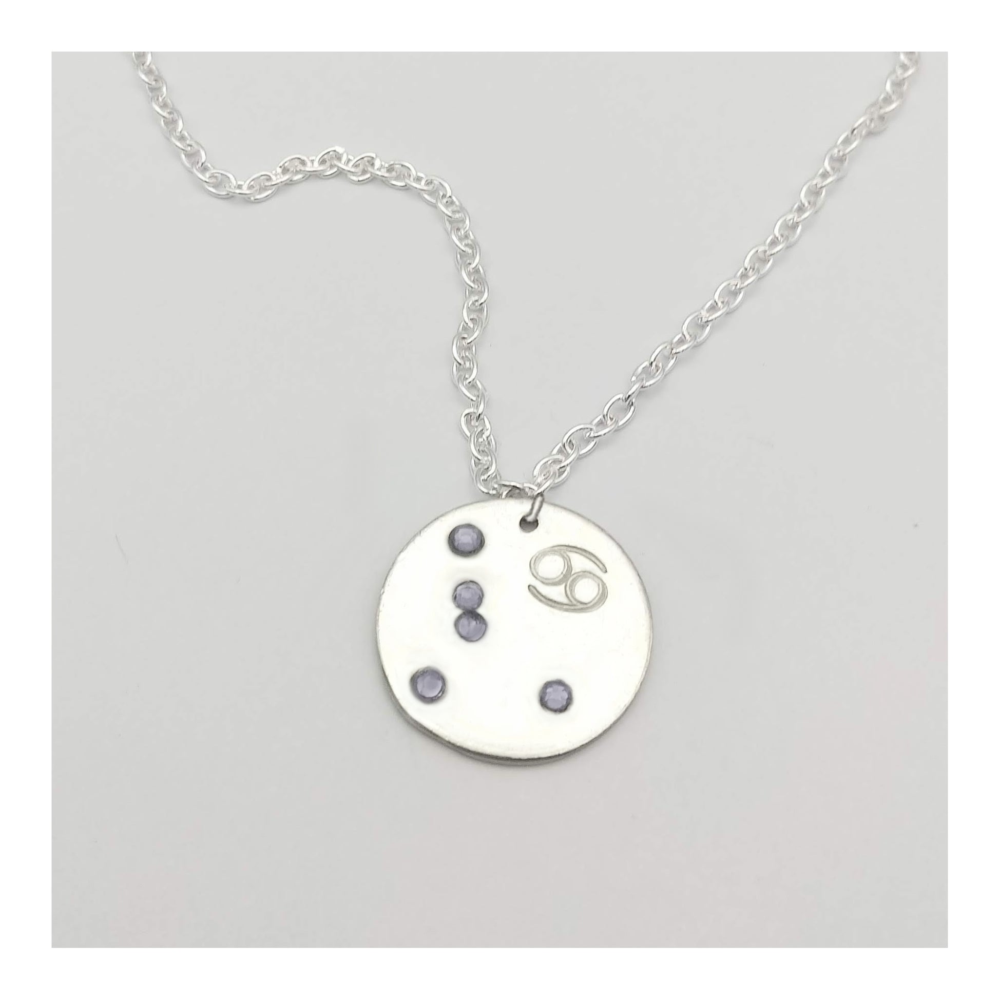 June Cancer necklace pendant with hand set alexandrite colored glass rhinestones forming the constellation of Cancer. Hand stamped with the zodiac symbol for Cancer, on a 3/4th of an inch silver toned circle hanging from a silver toned chain.