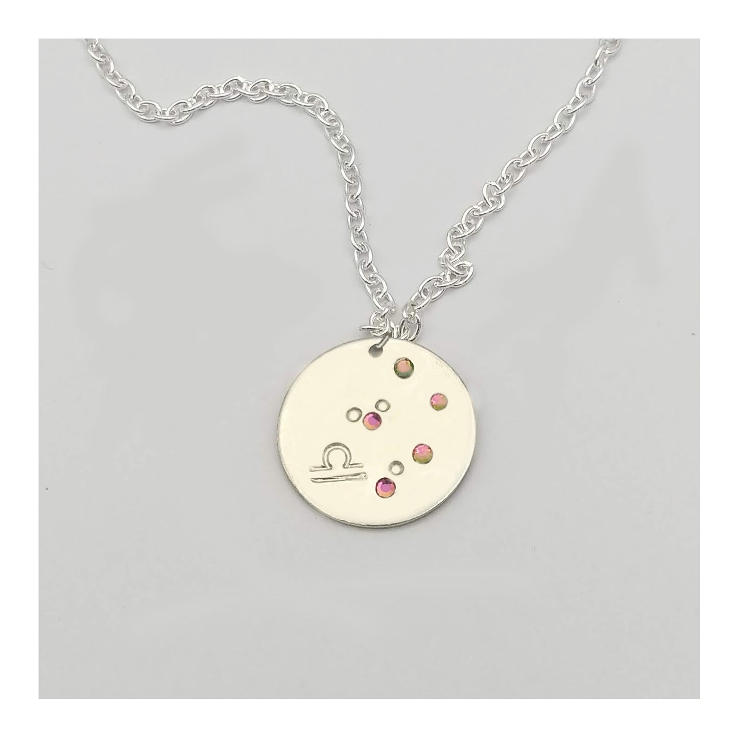 October Libra necklace pendant with hand set tourmaline colored glass rhinestones forming the constellation of Libra. Hand stamped with the zodiac symbol for Libra, on a 3/4th of an inch silver toned circle hanging from a silver toned chain.