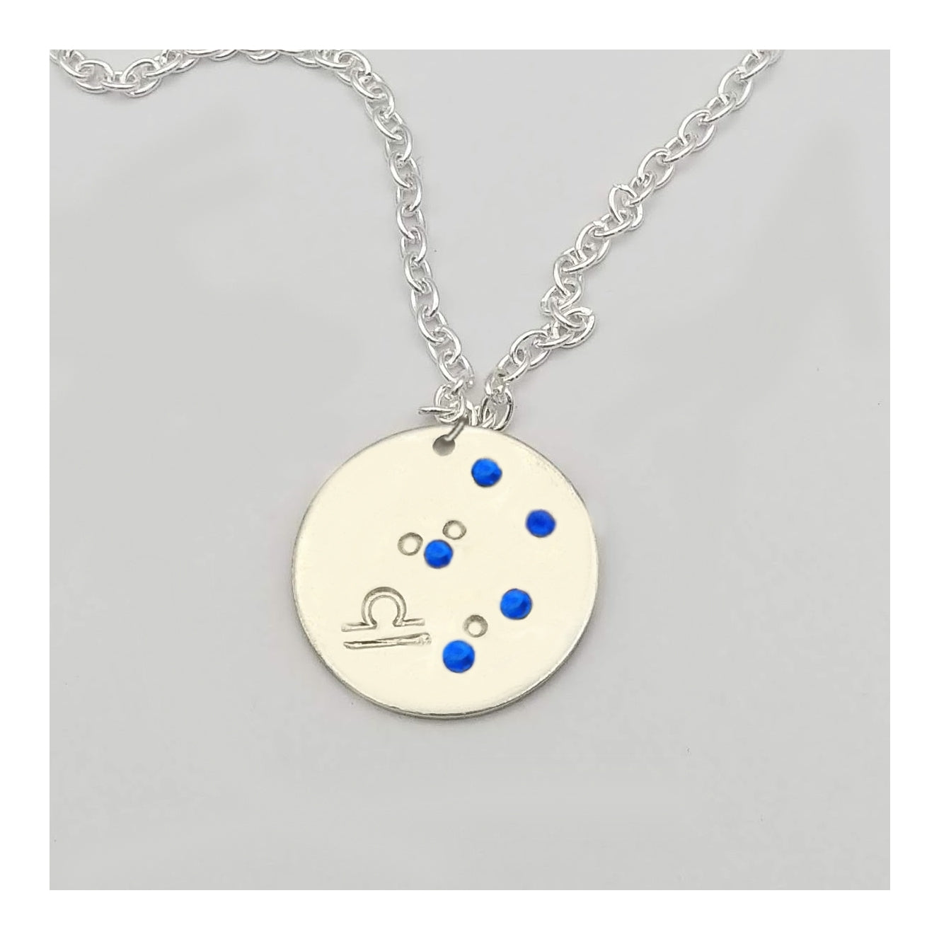 September Libra necklace pendant with hand set sapphire colored glass rhinestones forming the constellation of Libra. Hand stamped with the zodiac symbol for Libra, on a 3/4th of an inch silver toned circle hanging from a silver toned chain.