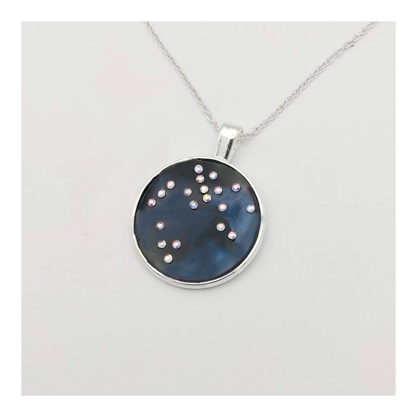 Dark blue with iridescent sparkling swirls of pinks, purples, silver, and monochromatic shades of blue enamel. With handset Aurora Borealis rhinestones to represent the star of the constellation of Sagittarius. The pendant is 1 inch silver toned with a 1/4th inch bail on a silver toned chain.