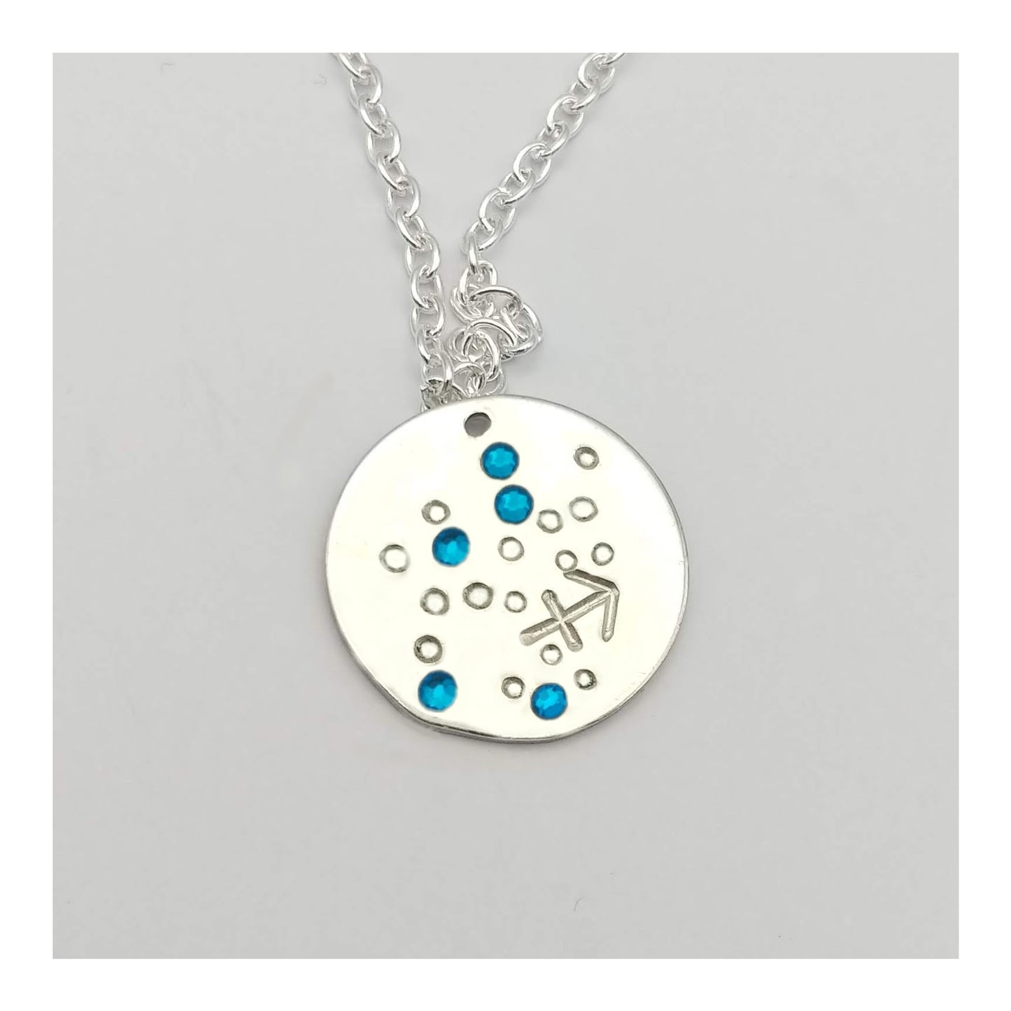 December Sagittarius necklace pendant with hand set tanzanite colored glass rhinestones forming the constellation of Sagittarius. Hand stamped with the zodiac symbol for Sagittarius, on a 3/4th of an inch silver toned circle hanging from a silver toned chain.