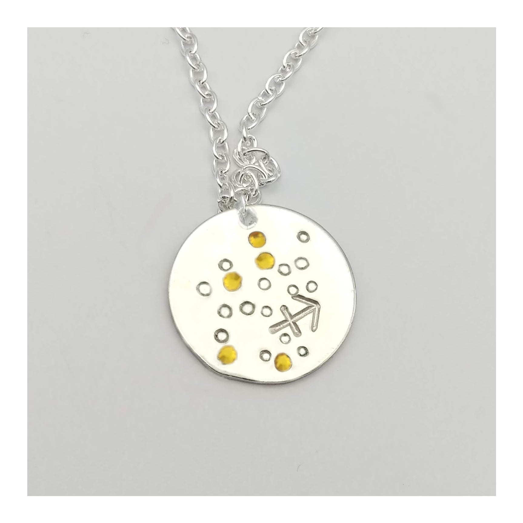 November Sagittarius necklace pendant with hand set citrine colored glass rhinestones forming the constellation of Sagittarius. Hand stamped with the zodiac symbol for Sagittarius, on a 3/4th of an inch silver toned circle hanging from a silver toned chain.