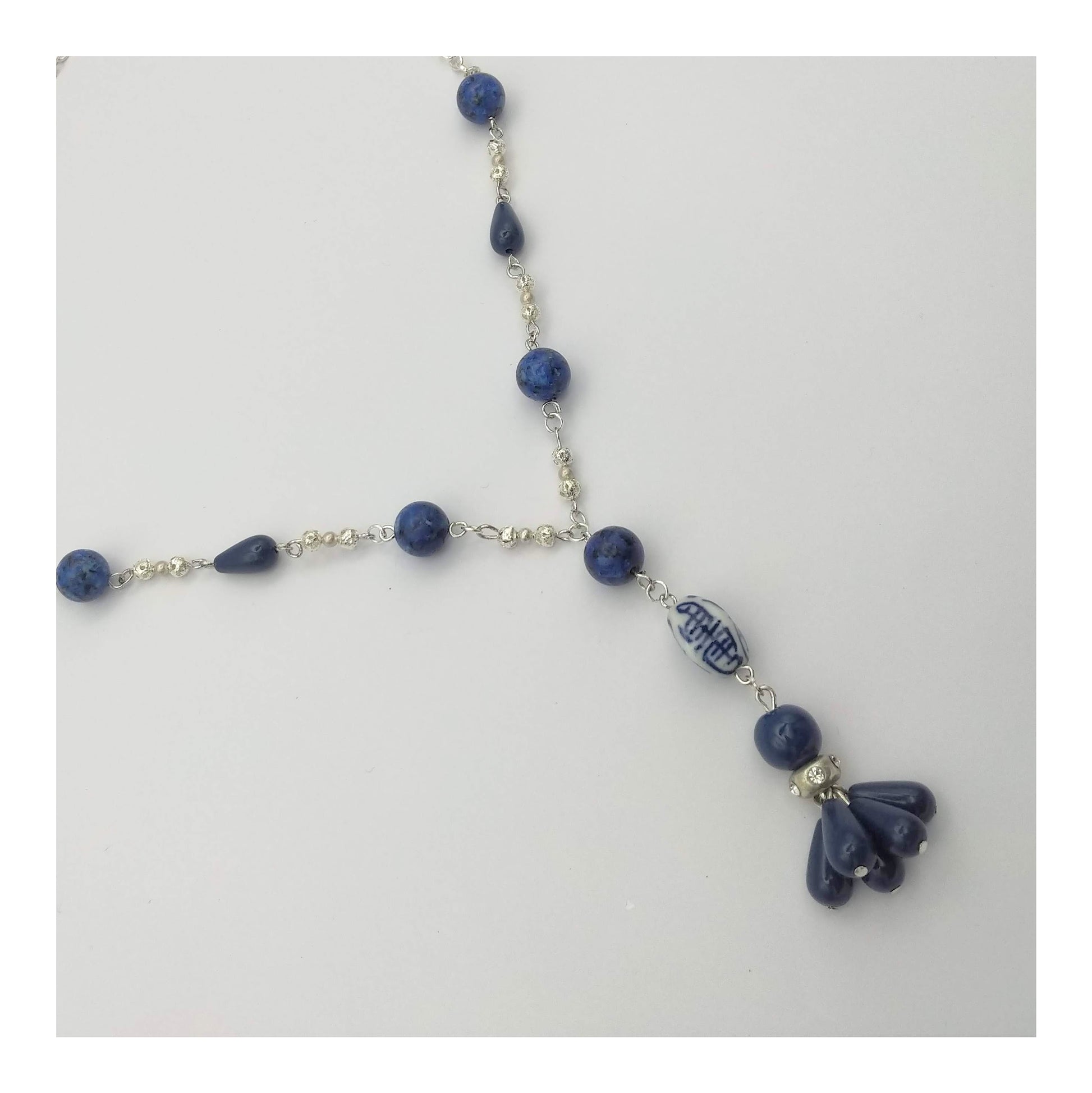 Silver toned necklace with round Lapis Lazuli beads accented with blue teardrop beads in a symmetrical pattern on a 20 inch long chain. A 3 inch drop designed with round lapis, oblong white and blue porcelain bead and round blue bead with matching teardrop tassels.   