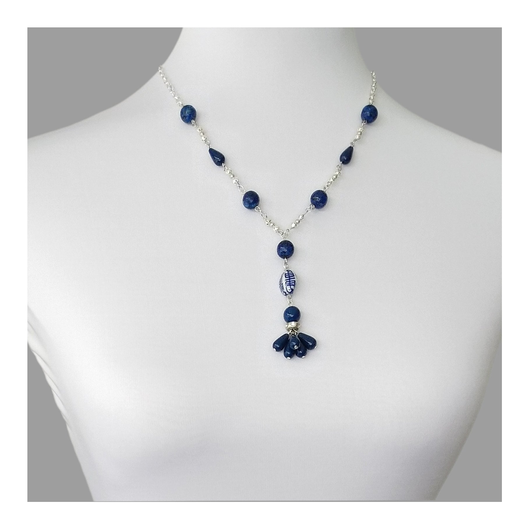 Silver toned necklace with round Lapis Lazuli beads accented with blue teardrop beads in a symmetrical pattern on a 20 inch long chain. A 3 inch drop designed with round lapis, oblong white and blue porcelain bead and round blue bead with matching teardrop tassels.   