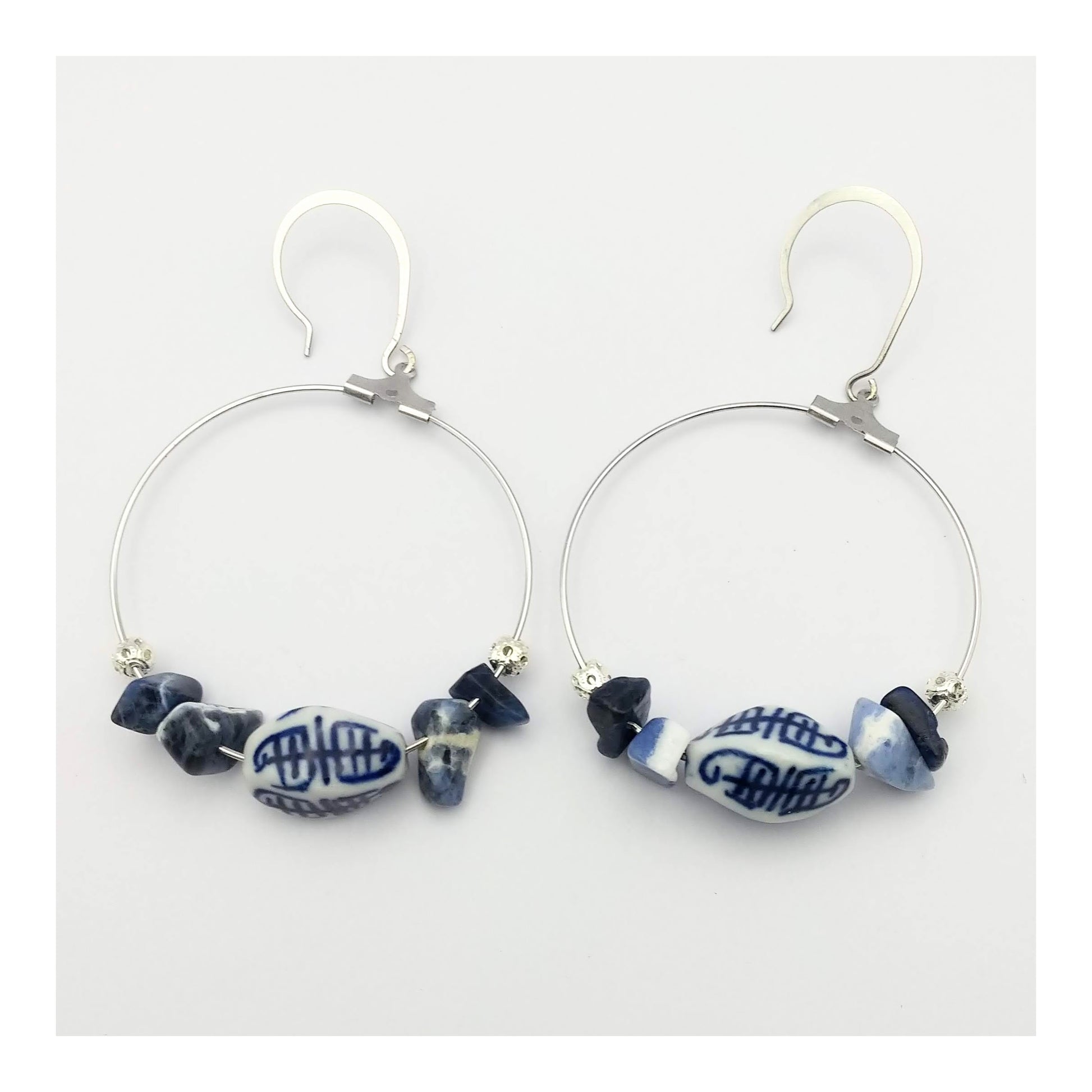 Sodalite chips on both sides of an oblong white with blue scribble detailed porcelain bead. A-lined on a silver toned 1 ½ inch diameter hoop attached to fish hooks to equal a 2 ½ inch drop.   